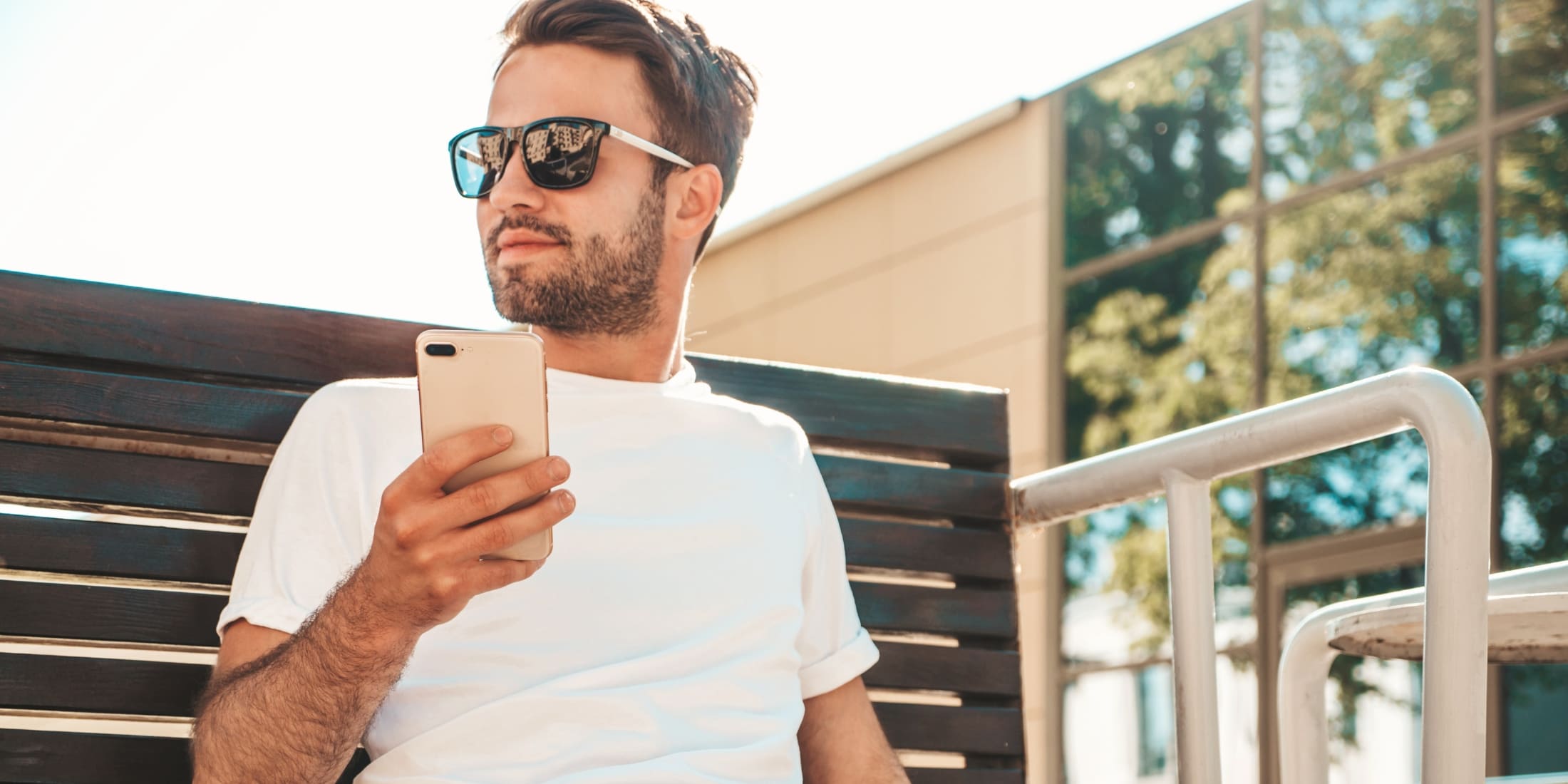 Man wearing sunglasses and a white t-shirt, sitting on a bench outdoors on a sunny day, holding a smartphone, and looking off into the distance. This image reflects the modern lifestyle and habits for men, emphasizing relaxation, mindfulness, and the importance of taking moments to reflect and recharge. The background includes modern glass buildings and reflections of trees, enhancing the serene and thoughtful atmosphere.