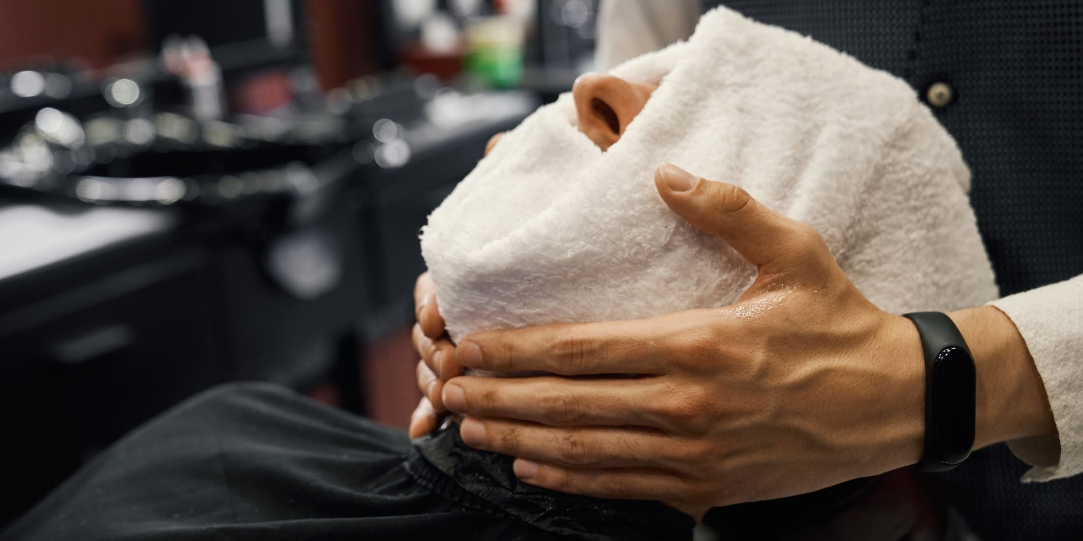 A man receiving a hot towel shave at a barbershop, highlighting the benefits of a hot towel shave. The barber is holding a warm towel wrapped around the man's face, showcasing the relaxing and skin-enhancing experience of a professional shave.