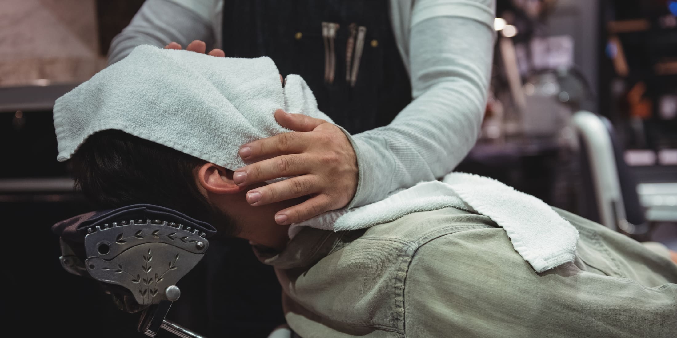 A man experiencing a hot towel shave at a barbershop, with a barber applying a warm towel to his face. This process highlights the benefits of a hot towel shave, including deep cleansing, exfoliation, and relaxation, for a luxurious grooming experience.