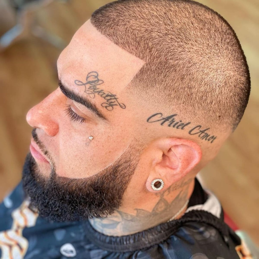 A close-up of a man with a precise haircut, featuring sharp lines along the hairline and a meticulously styled beard. His tattoos add a bold, personal touch, while his clean-cut style highlights his facial features. The look is completed with a stud earring and a small facial piercing, adding to the edgy, polished appearance.