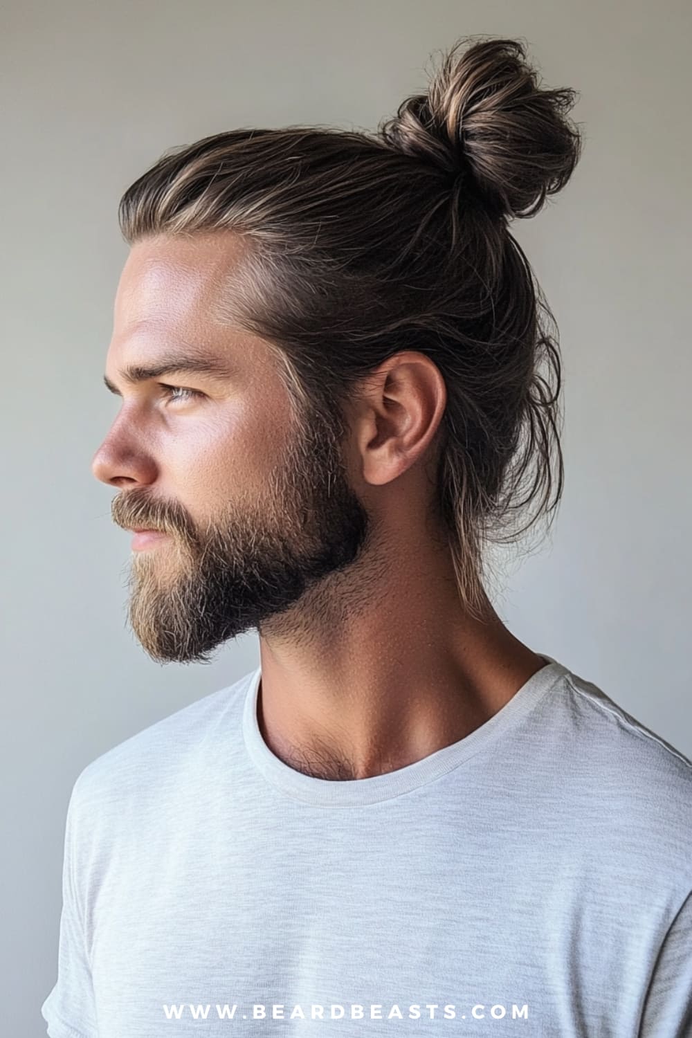 A side profile of a man showcasing a classic man bun hairstyle. His dark hair is smoothly pulled back into a neat bun at the crown of his head, exuding a clean and timeless look. The man has a well-groomed beard that complements the sleek hairstyle, adding to his refined yet casual appearance. He is wearing a simple white t-shirt, which further emphasizes the effortless style of the classic man bun. The plain, neutral background keeps the focus on the man’s hairstyle and overall polished look.