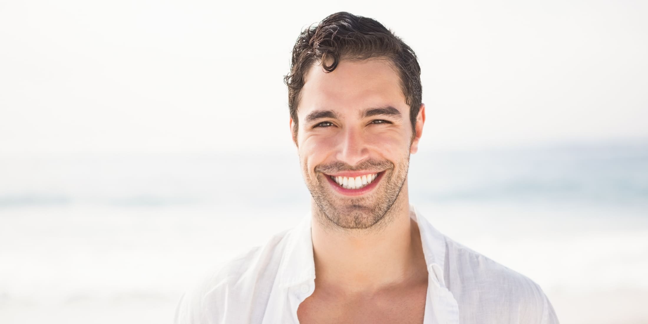 Close-up of a young man with a light stubble beard, smiling confidently at the camera on a bright beach day. The natural sunlight highlights his healthy skin and well-groomed beard, showcasing the early stages of beard growth. Perfect representation of when to start using beard oil for optimal beard care.
