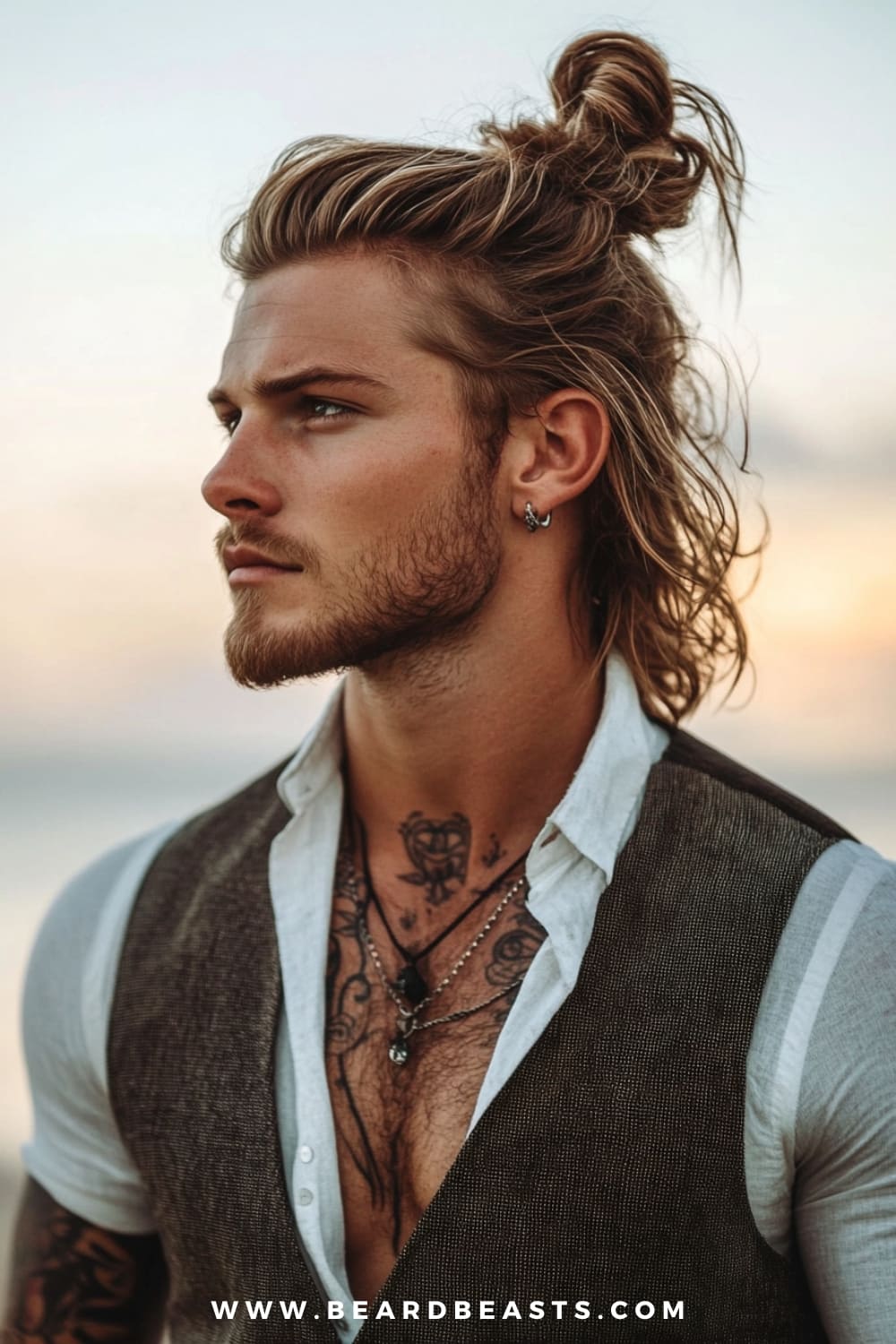 A striking image of a man showcasing one of the popular man bun hairstyles—the half-up, half-down man bun. His long, wavy blonde hair is partially pulled back into a casual bun, while the rest flows freely. He has a well-groomed beard, adding to the overall rugged yet stylish vibe. Dressed in a white shirt and a brown vest, his look is completed with visible tattoos and earrings, enhancing the edgy appeal. The background, softly blurred with a sunset glow, creates a warm, serene atmosphere, perfectly complementing the man bun style.