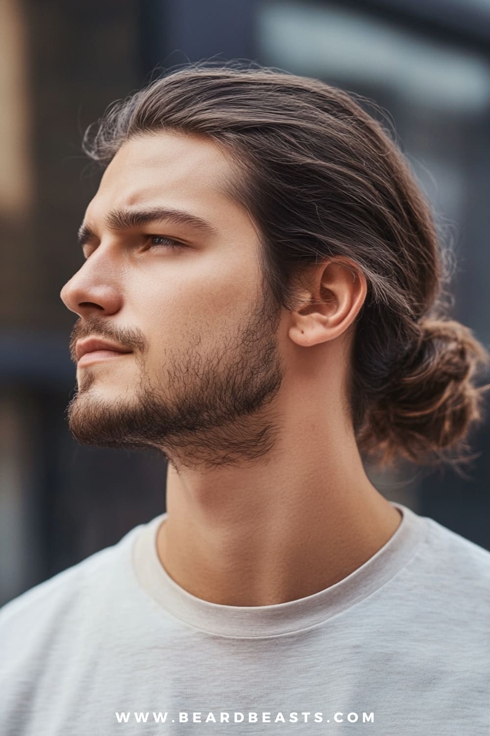 A side profile of a man with a relaxed, low bun hairstyle. His dark, slightly wavy hair is casually pulled back into a bun that sits at the nape of his neck, giving off a laid-back and effortless vibe. The man has a light, natural beard that complements his soft, contemplative expression. He is wearing a simple white t-shirt, adding to the overall minimalistic and casual look. The blurred background suggests an outdoor setting, subtly highlighting the man's calm and serene presence.