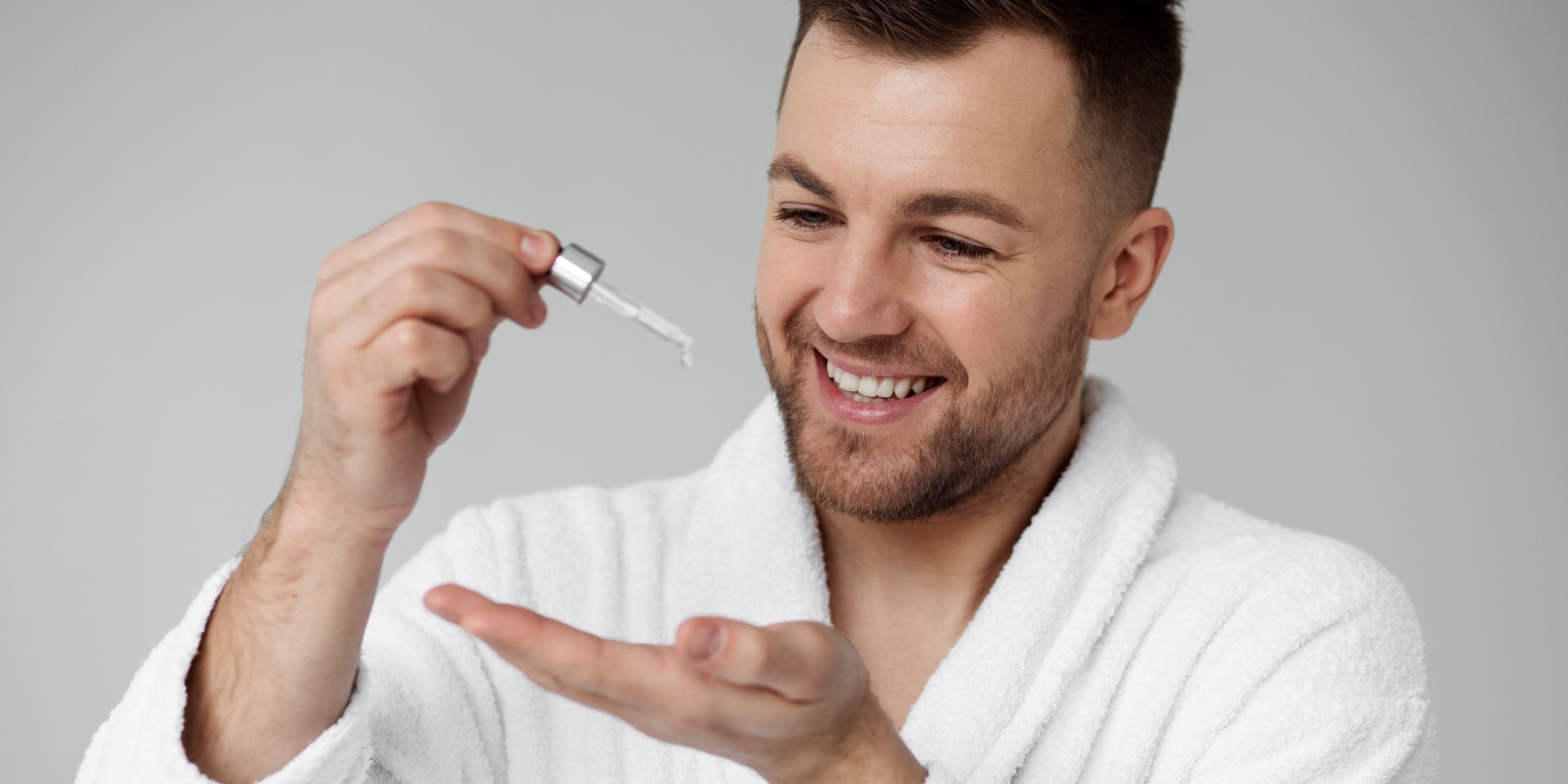 Smiling man in a white robe applying pre-shave oil to his hand, demonstrating what is pre shave oil and how to use it for a smoother, more comfortable shaving experience.