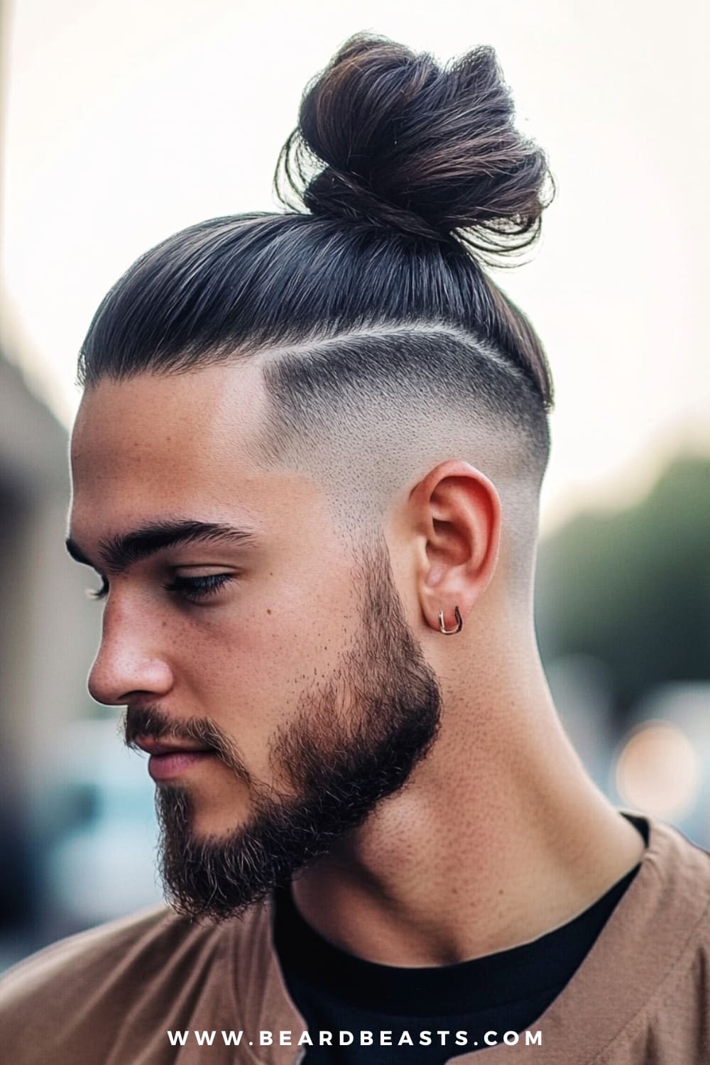 A stylish image of a man featuring an undercut man bun hairstyle. His dark, sleek hair is pulled back into a high, tight bun on top, while the sides are shaved close to the scalp, creating a sharp contrast that highlights the undercut. The clean lines of the fade enhance the modern, edgy look. The man has a well-groomed beard and wears a small hoop earring, adding to the overall stylish and confident appearance. The background is softly blurred, keeping the focus on the striking man bun hairstyle.