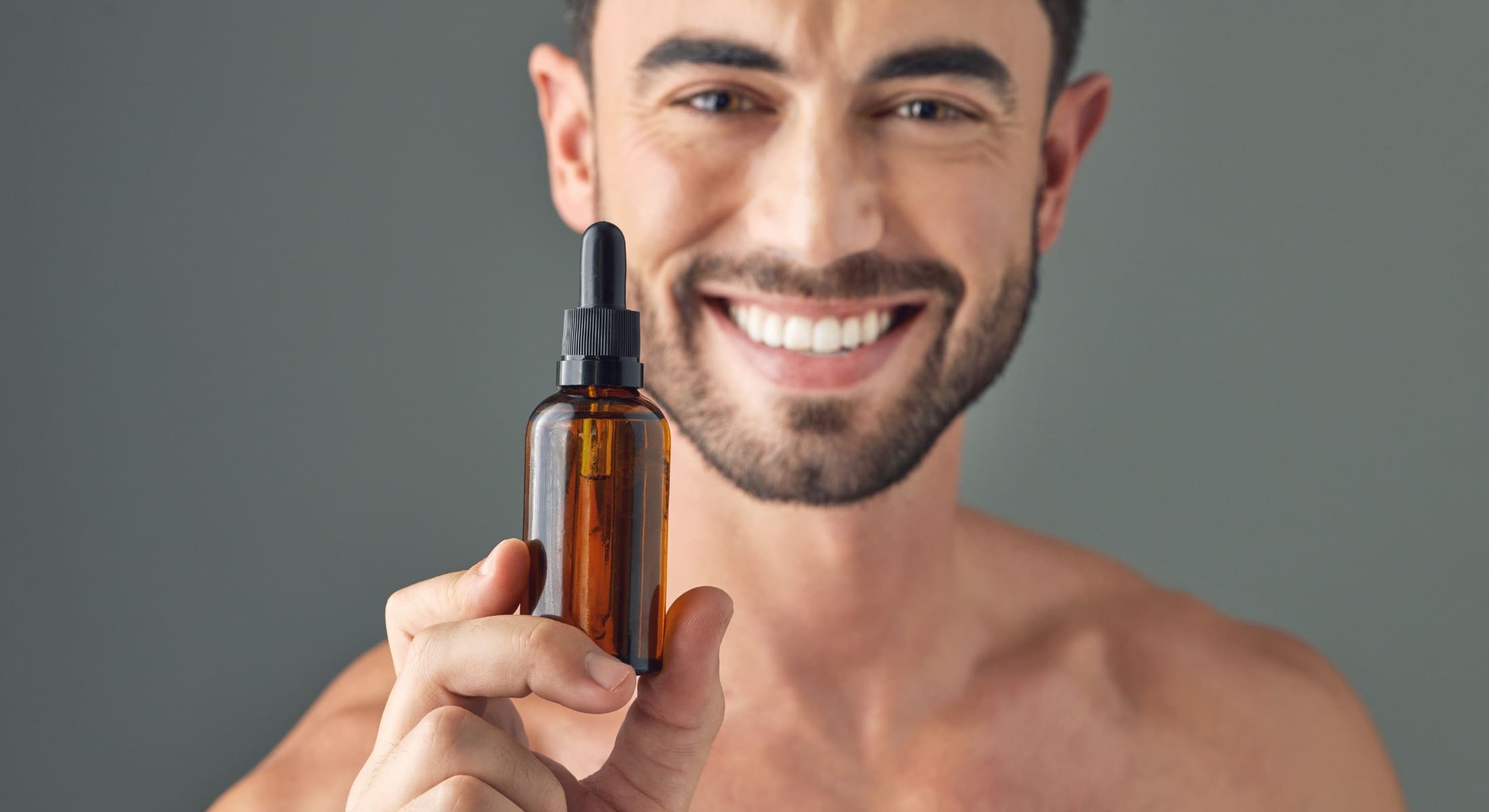 Smiling man holding a bottle of beard oil, emphasizing the benefits of using beard oil every day for a well-groomed and healthy beard.