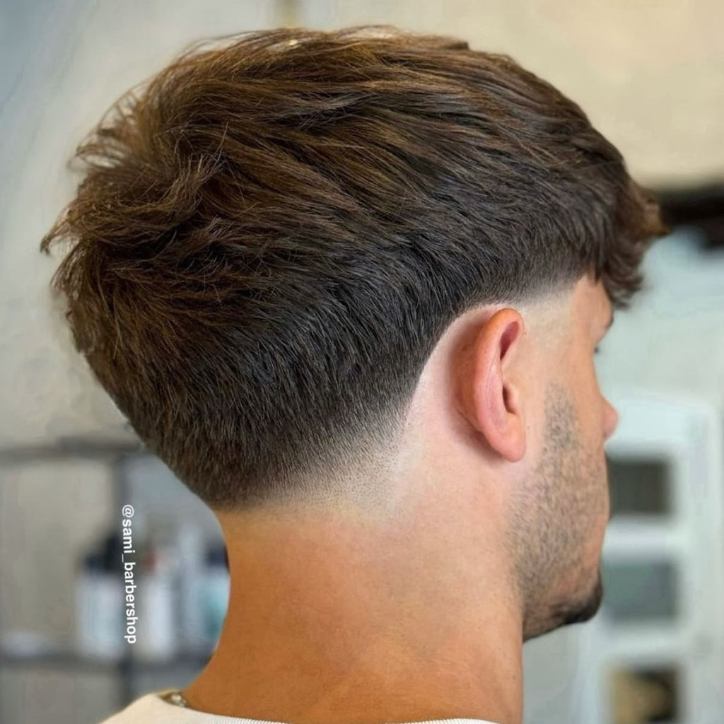 A back view of a man with a classic taper cut, featuring a soft, blended gradient from longer hair on top to a shorter length at the nape of the neck. The hair is slightly textured, giving a natural and full appearance, while the taper creates a clean and structured outline around the ears and neckline. This haircut has a timeless appeal, providing a neat and balanced look suitable for various styles and occasions.