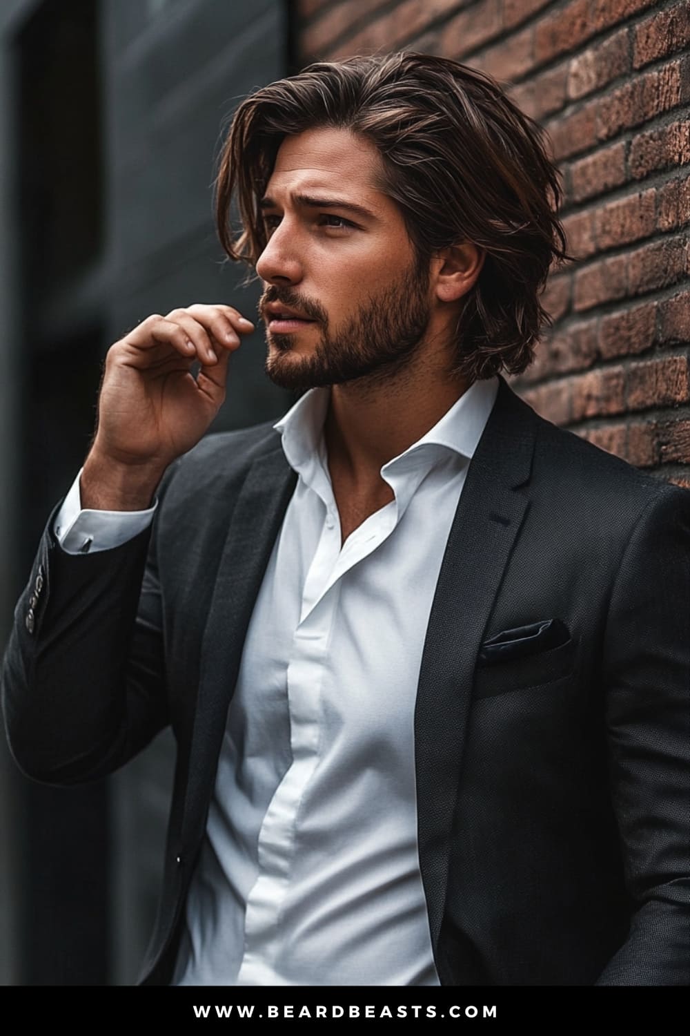 Handsome man with long, layered hair and a neatly trimmed beard, dressed in a sharp suit, leaning against a brick wall. This look perfectly captures a stylish and modern example of men's hairstyles for long hair, combining elegance with a relaxed, confident vibe.