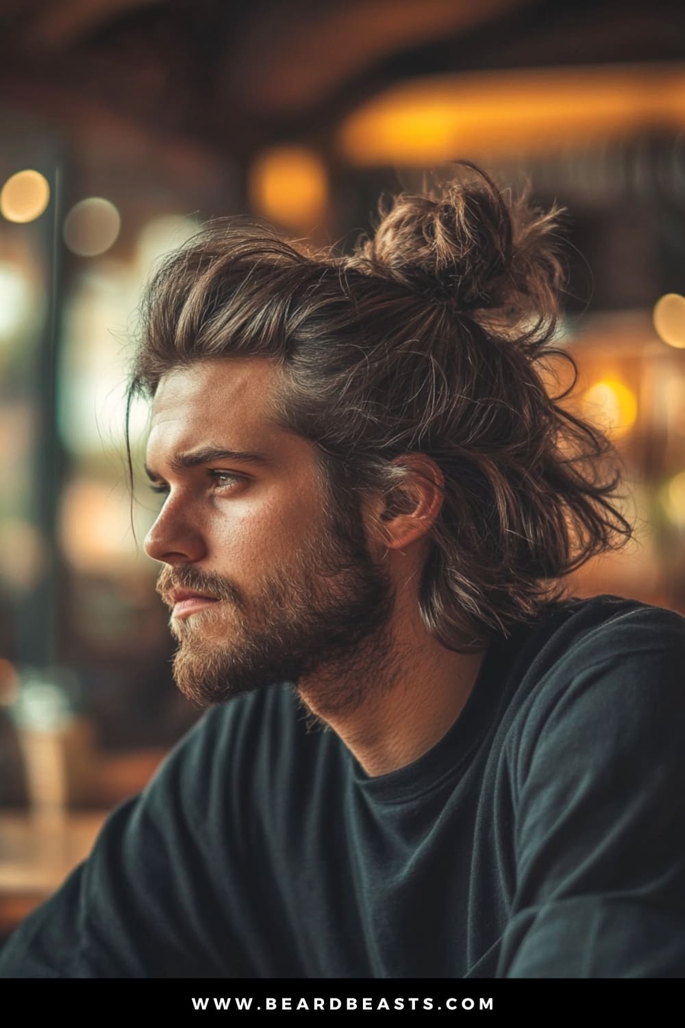 Man with a casual yet stylish messy bun and a well-groomed beard, sitting in a cozy café setting. The relaxed, tousled look exemplifies the perfect men's hairstyle for long hair, combining effortless cool with a laid-back, modern aesthetic.