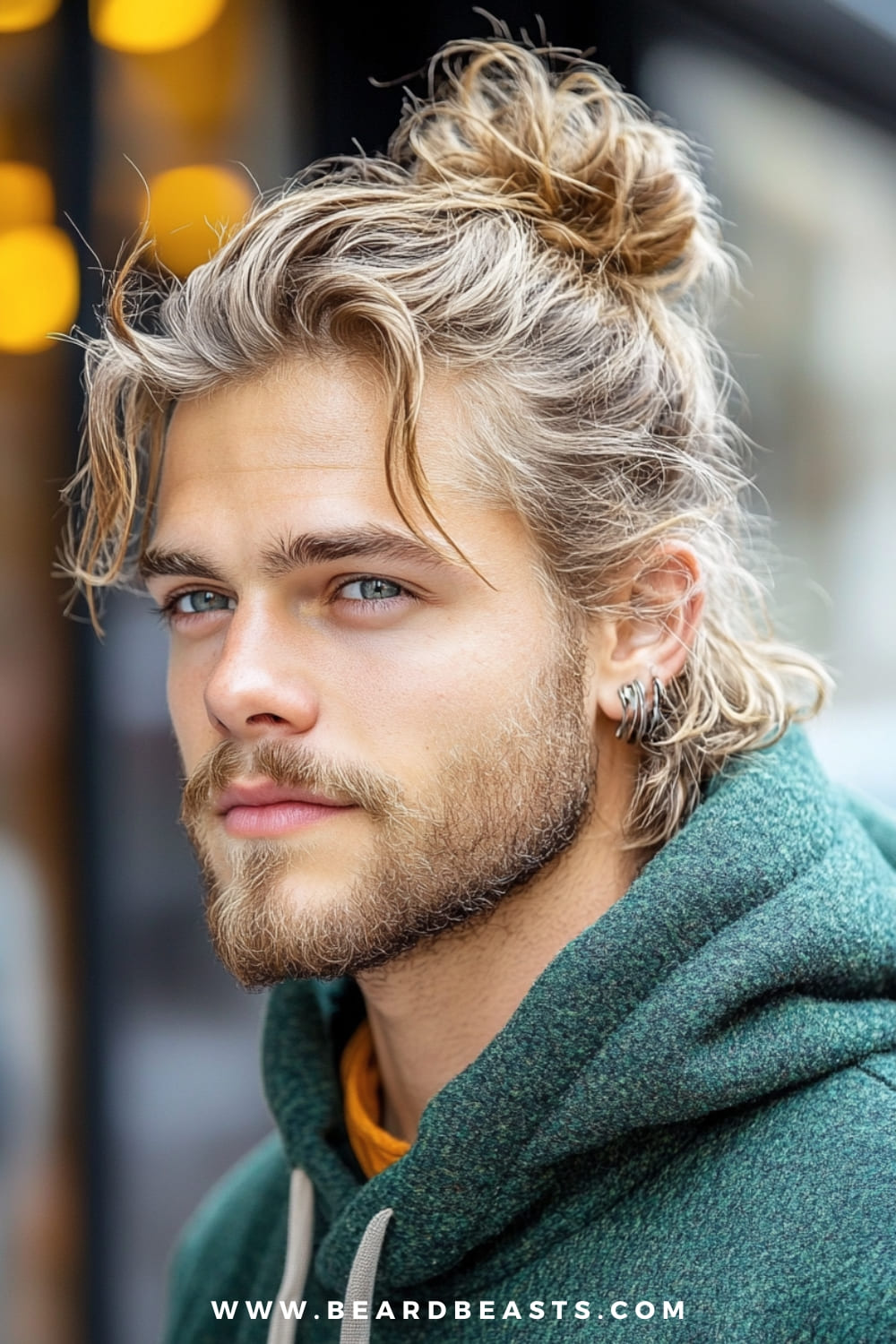 A captivating image of a man sporting a messy man bun hairstyle. His tousled, blonde hair is loosely pulled back into a casual bun, with a few strands framing his face for a relaxed, effortlessly cool look. He has a well-groomed beard and is wearing a green hoodie, adding to the laid-back vibe of the style. The man’s piercing blue eyes and subtle earrings enhance his stylish appearance. The blurred background, featuring warm tones, keeps the focus on the man and his trendy man bun hairstyle.