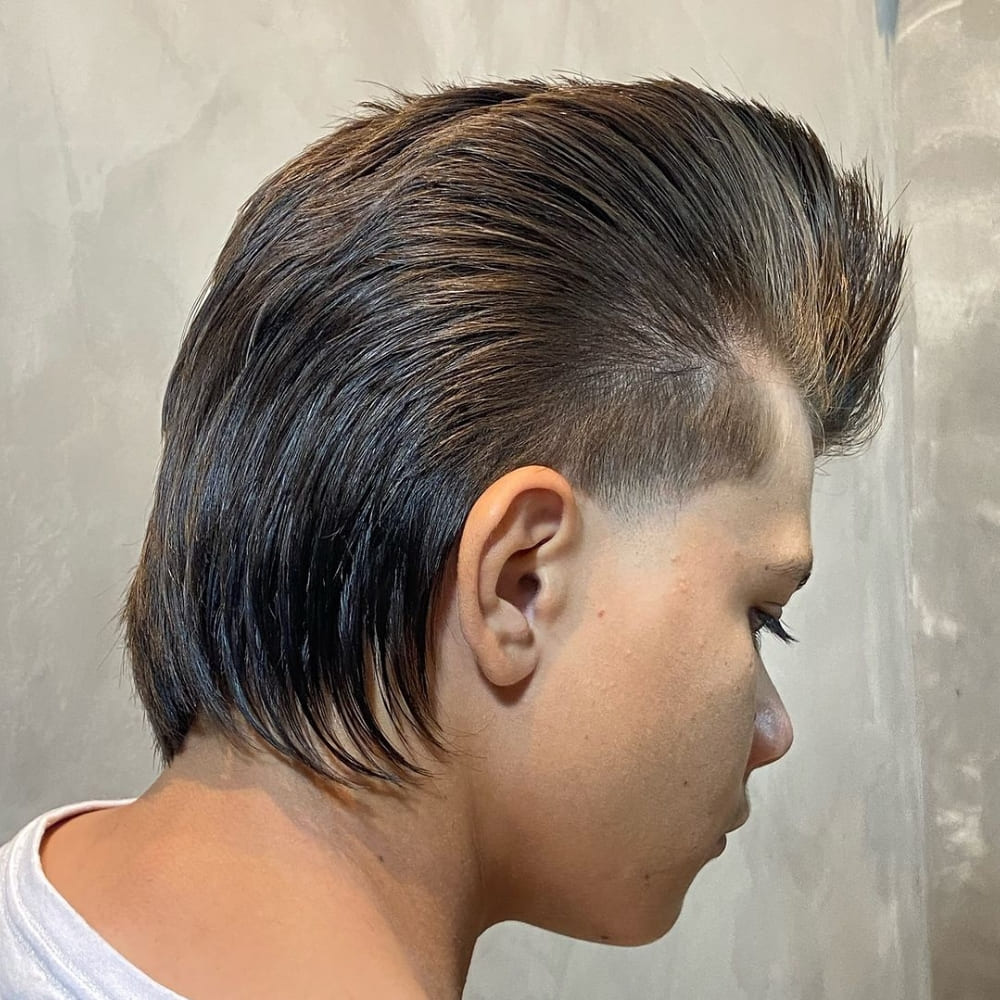 A young man is showcasing a distinctive mullet pompadour haircut. The top is styled high and slicked back, creating a voluminous pompadour effect. The sides are neatly tapered and faded, providing a clean and sharp contrast. The back features a classic mullet style, with longer hair flowing down the neck, adding a retro yet modern twist to the overall look. The man’s expression is focused, highlighting the bold and edgy nature of his unique hairstyle. The background is neutral, allowing the haircut to stand out prominently. A network error occurred. Please check your connection and try again. If this issue persists please contact us through our help center at help.openai.com.