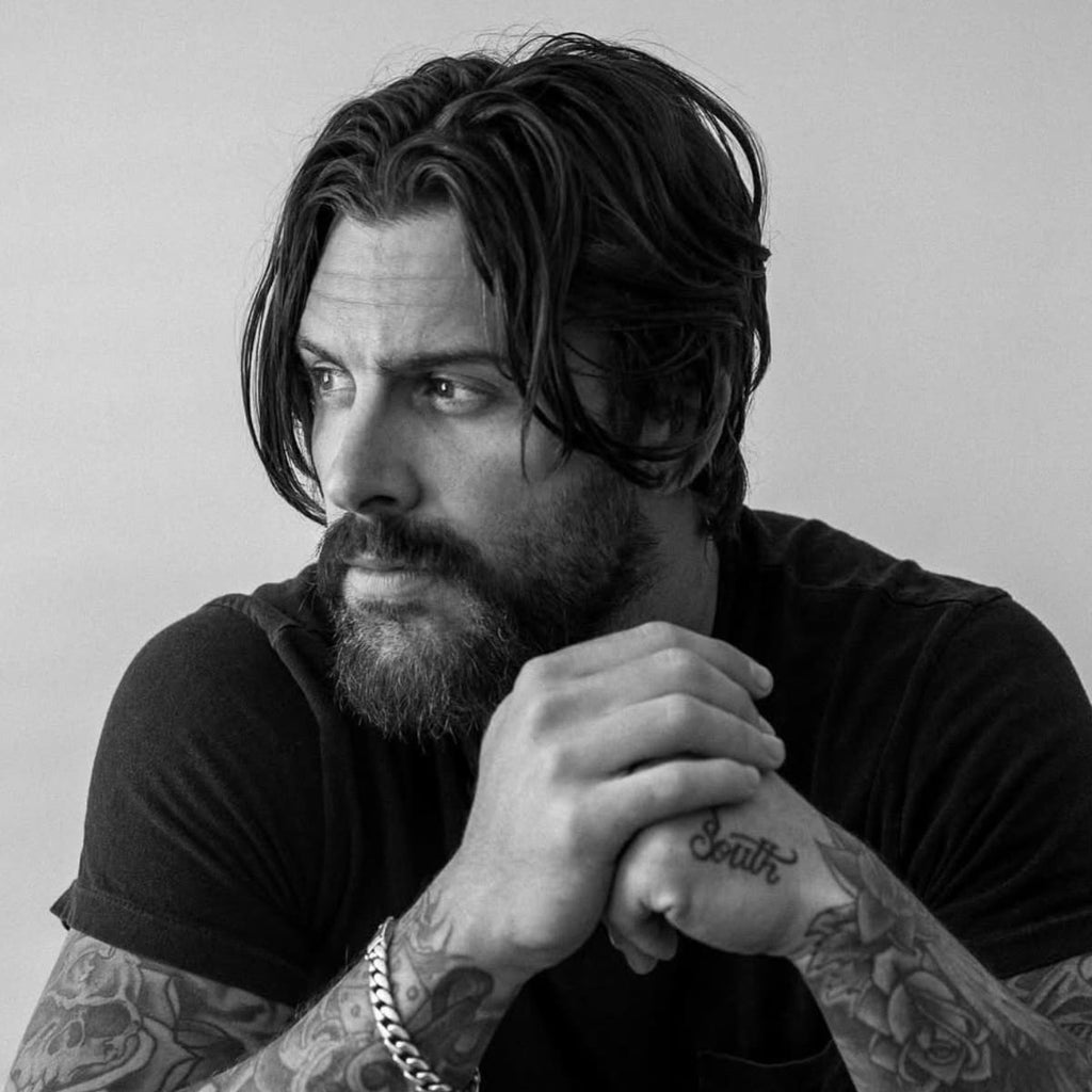 A contemplative black and white portrait of a man with rugged facial hair and tousled long hair. He gazes thoughtfully to the side, his intense eyes and furrowed brow adding depth to his expression. His facial hair is thick and slightly unkempt, framing his face and complementing his casual black t-shirt. Tattooed arms folded, he exudes a raw, introspective aura against a neutral background. The image captures a moody, artistic vibe, highlighting his striking facial features and casual, yet deliberate style.