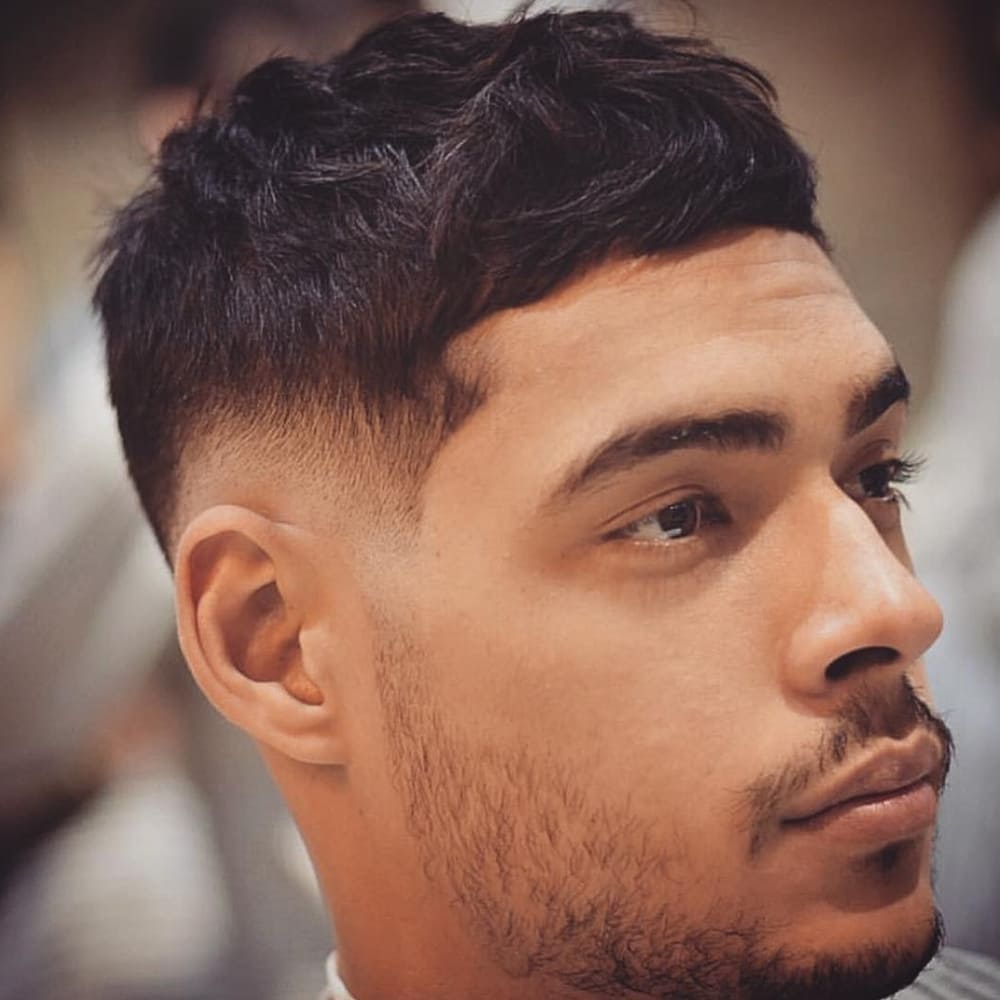 A young man with a textured crop low fade haircut is shown in the image. His hair features a neatly styled, textured top with subtle layers that add depth and movement. The sides and back are smoothly faded low, creating a gentle transition from the longer hair on top to the shorter hair around the ears and neckline. This low fade enhances the overall clean and modern look. The man has a light beard, complementing his sharp hairstyle and adding to his polished appearance. The background is blurred, keeping the focus on his well-executed haircut and facial features.