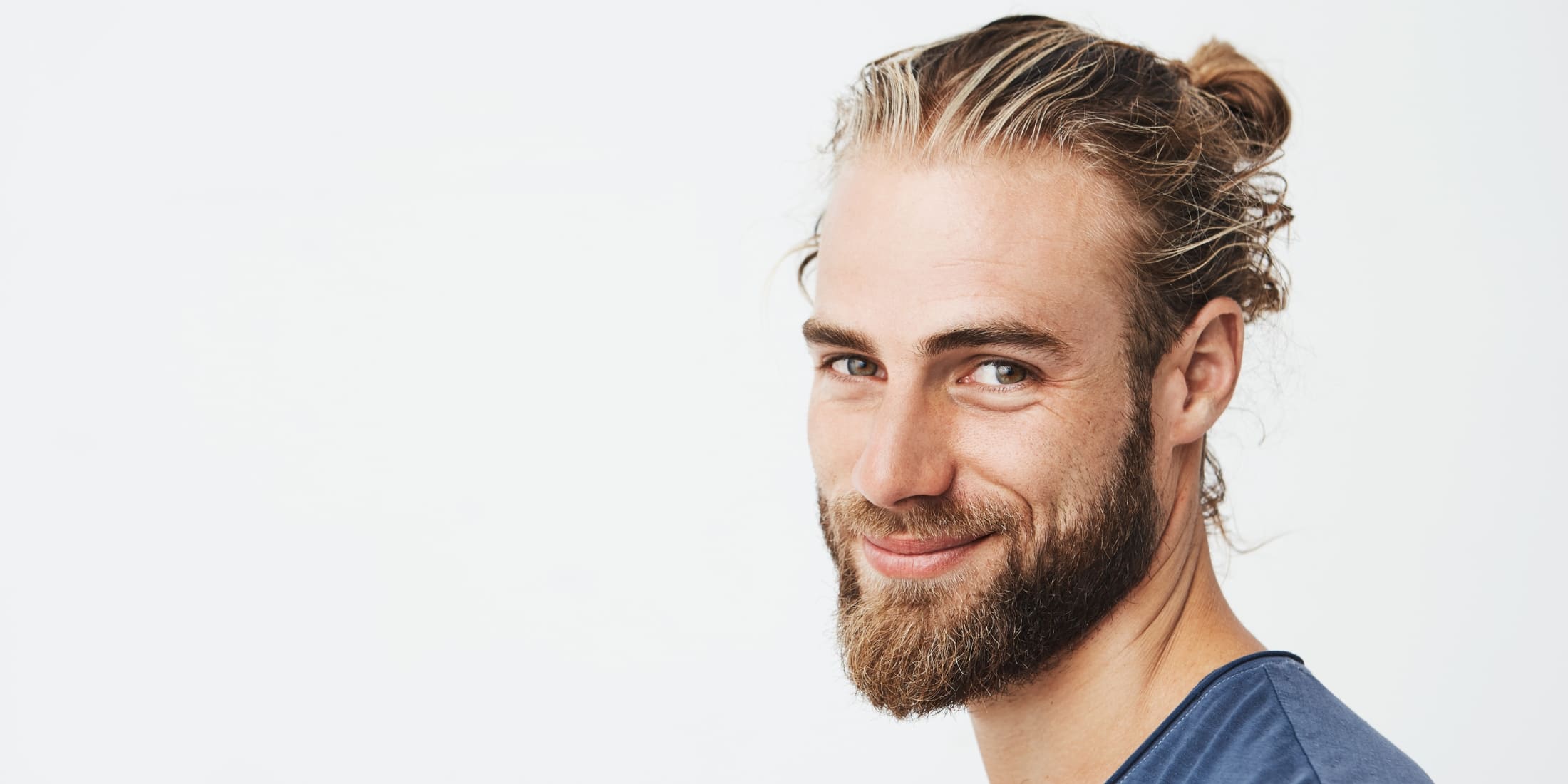 Smiling man with a well-groomed short beard and a tied-back man bun hairstyle, wearing a blue shirt. The image illustrates the results of proper short beard grooming, including techniques for trimming, shaping, and maintaining a healthy beard.
